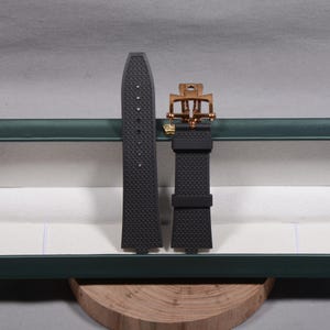 May include: Black watch strap with a textured pattern and a gold-coloured buckle. The strap is made of a flexible material and is designed to be durable. The buckle has a unique design. The watch strap is displayed on a wooden surface.