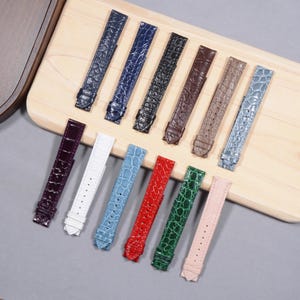 Könnte beinhalten: Eine Sammlung von Armbändern in verschiedenen Farben, darunter Blau, Schwarz, Braun und Rosa. Die Armbänder haben ein strukturiertes, krokodilartiges Muster. Sie sind auf einer hellen Holzoberfläche angeordnet.