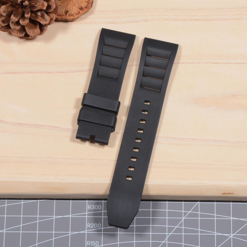 Op de afbeelding: Twee zwarte horlogebandjes worden getoond op een licht houten oppervlak. De bandjes zijn gemaakt van een flexibel materiaal, met een textuurontwerp. E&eacute;n bandje heeft een gesp en de andere heeft verstelgaten. De bandjes zijn ontworpen voor een horloge.