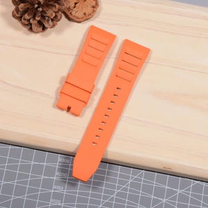 Op de afbeelding: Twee feloranje horlogebandjes worden gepresenteerd op een licht houten oppervlak. De bandjes zijn gemaakt van een flexibel materiaal, met een reeks rechthoekige inkepingen aan &eacute;&eacute;n uiteinde en een reeks gaten aan het andere. De bandjes zijn ontworpen voor een horloge.