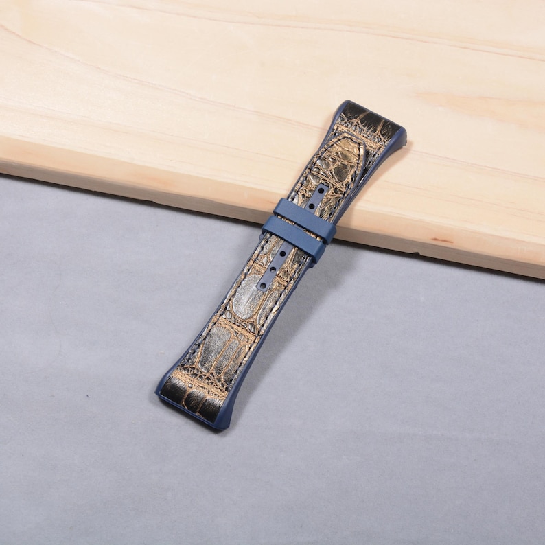 K&ouml;nnte beinhalten: Ein blau-goldenes Uhrenarmband mit Krokodilleder-Muster. Das Armband hat einen blauen Verschluss und besteht aus einem strukturierten Material. Das Uhrenarmband liegt auf einer hellen Holzoberfl&auml;che.