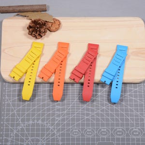 Op de afbeelding: Vier kleurrijke horlogebandjes in geel, oranje, rood en blauw. Elk bandje is gemaakt van flexibel materiaal en heeft verstelgaten. De bandjes zijn gerangschikt op een houten snijplank.