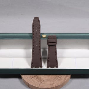 Puede incluir: Una correa de reloj marr&oacute;n con una superficie texturizada. La correa est&aacute; hecha de un material duradero y presenta costuras a lo largo de los bordes. La correa se muestra dentro de una caja verde con un emblema de corona dorada.