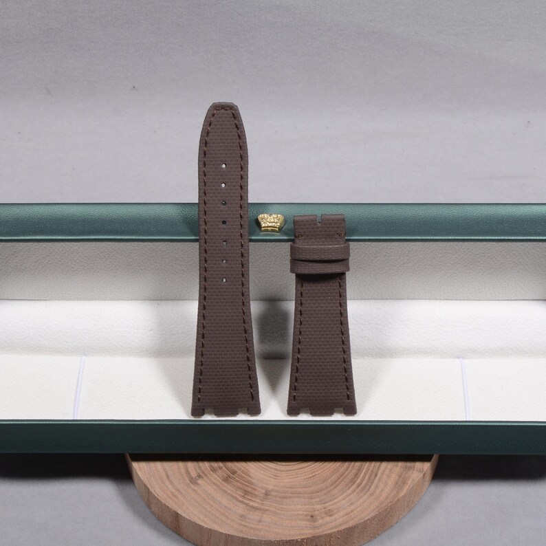 Puede incluir: Una correa de reloj marr&oacute;n con una superficie texturizada. La correa est&aacute; hecha de un material duradero y presenta costuras a lo largo de los bordes. La correa se muestra dentro de una caja verde con un emblema de corona dorada.