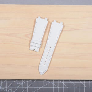May include: Two white leather watch straps with contrasting white stitching. One strap has a buckle, and the other has several holes for adjustment. The straps are displayed on a light wooden surface.