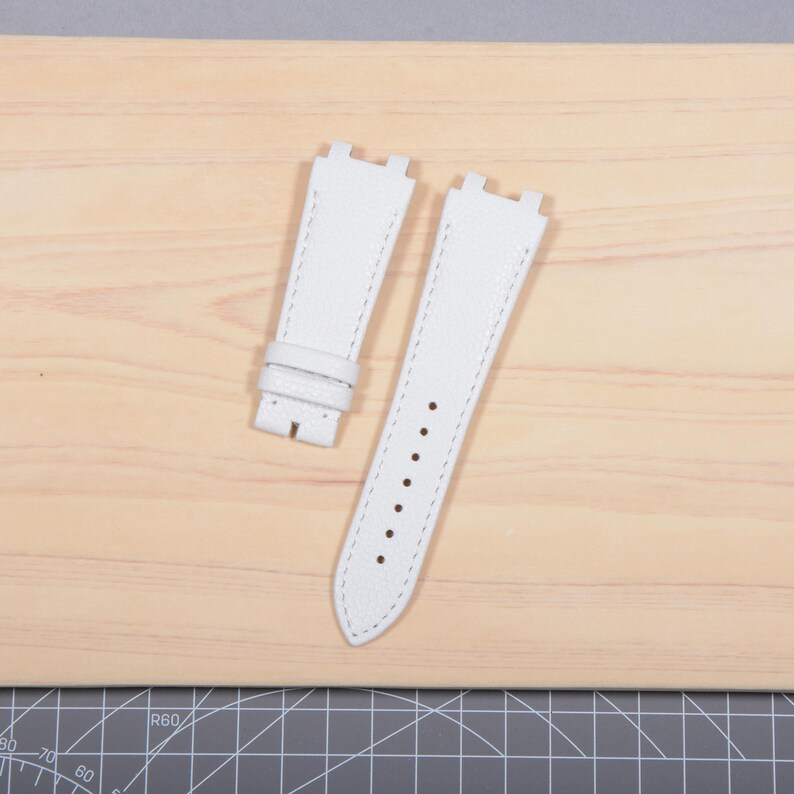 May include: Two white leather watch straps with contrasting white stitching. One strap has a buckle, and the other has several holes for adjustment. The straps are displayed on a light wooden surface.