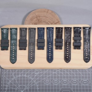 May include: A collection of eight watch straps in various colors, including green, blue, gray, and black. The straps are made of a textured material, possibly leather, and are displayed on a wooden board. Each strap has a black buckle.