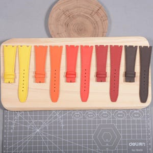Puede incluir: Una colecci&oacute;n de correas de reloj coloridas en tonos amarillo, naranja, rojo y marr&oacute;n. Las correas se exhiben en una tabla de madera, mostrando sus diversos colores y dise&ntilde;os. Cada correa tiene m&uacute;ltiples agujeros para ajuste.