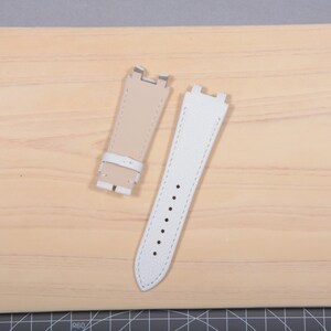 May include: Two watch straps. One is off-white with a buckle, the other is white with several holes. Both straps have white stitching and are made of leather. The straps are on a light brown wooden surface.