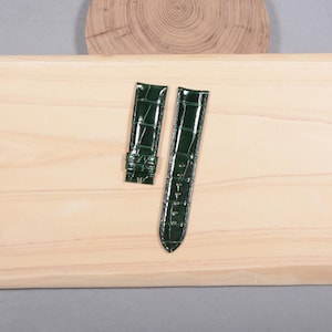 Puede incluir: Dos correas de reloj verde oscuro con textura de piel de cocodrilo se muestran sobre una superficie de madera clara. Las correas están hechas de un material brillante y están diseñadas para sujetarse a una esfera de reloj. Las correas son nuevas y sin usar.