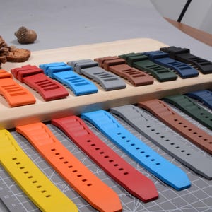 Op de afbeelding: Een verzameling kleurrijke horlogebandjes in verschillende tinten, waaronder geel, oranje, rood, blauw, grijs, bruin, groen en zwart. De bandjes zijn gemaakt van flexibel materiaal en hebben een reeks gaten voor aanpassing. Ze worden gepresenteerd op een houten oppervlak.