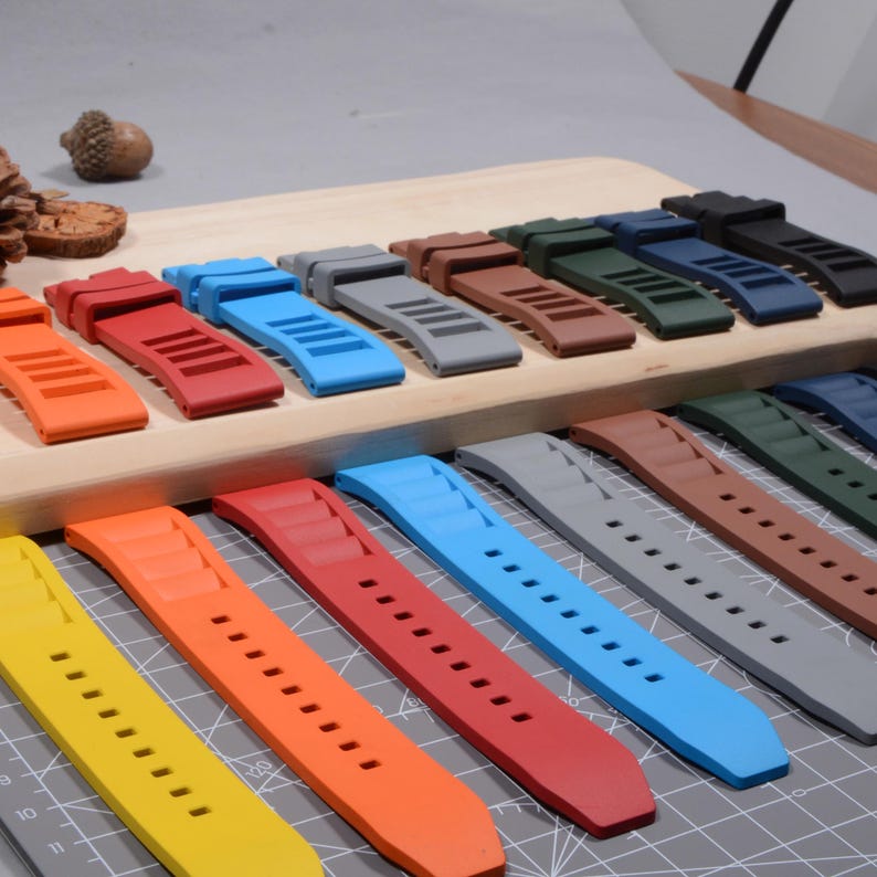 Op de afbeelding: Een verzameling kleurrijke horlogebandjes in verschillende tinten, waaronder geel, oranje, rood, blauw, grijs, bruin, groen en zwart. De bandjes zijn gemaakt van flexibel materiaal en hebben een reeks gaten voor aanpassing. Ze worden gepresenteerd op een houten oppervlak.