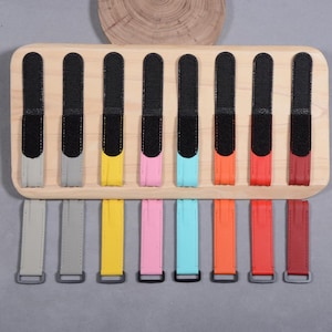 May include: Eight watch straps arranged in a row on a light wood display board. Each strap features a black hook-and-loop fastener at the top and a colored textured band at the bottom, available in gray, yellow, pink, light blue, orange, red, and dark red.