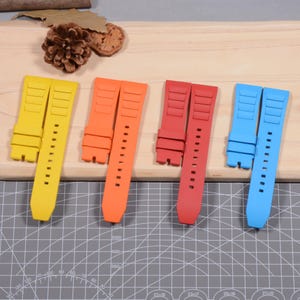 Op de afbeelding: Vier kleurrijke horlogebandjes in geel, oranje, rood en blauw. Elk bandje is gemaakt van een flexibel materiaal met verstelgaten en een gestructureerd ontwerp. De bandjes worden gepresenteerd op een houten oppervlak.