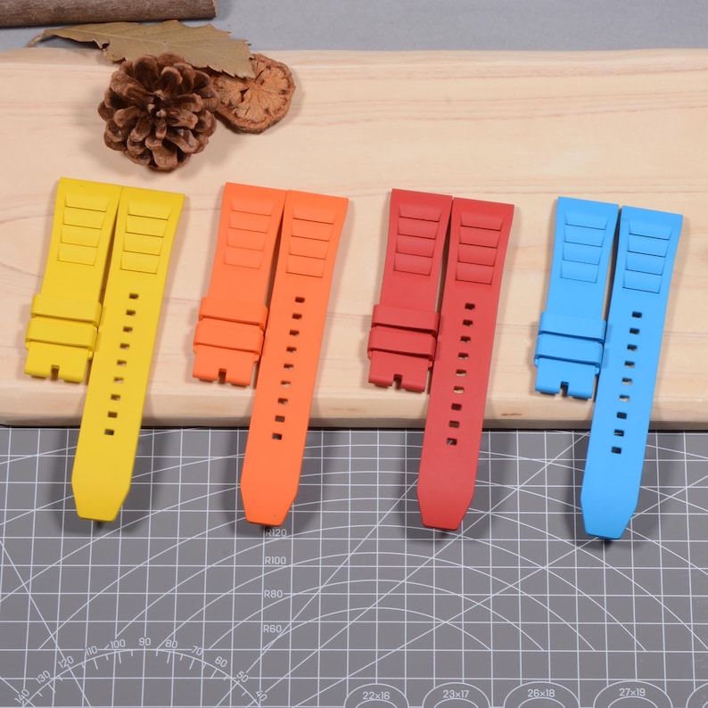 Op de afbeelding: Vier kleurrijke horlogebandjes in geel, oranje, rood en blauw. Elk bandje is gemaakt van een flexibel materiaal met verstelgaten en een gestructureerd ontwerp. De bandjes worden gepresenteerd op een houten oppervlak.