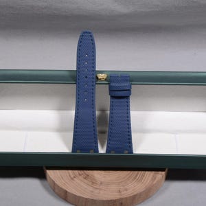 Puede incluir: Un par de correas de reloj azul marino con costuras blancas, presentadas en una caja verde abierta con un emblema dorado. Las correas est&aacute;n hechas de un material texturizado y descansan sobre una base de madera.