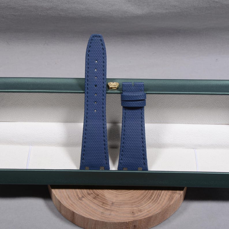 Puede incluir: Un par de correas de reloj azul marino con costuras blancas, presentadas en una caja verde abierta con un emblema dorado. Las correas est&aacute;n hechas de un material texturizado y descansan sobre una base de madera.