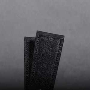 May include: Close-up of two black leather straps, possibly for a watch or bag. The straps have a textured surface and are stitched along the edges. The background is a solid gray.