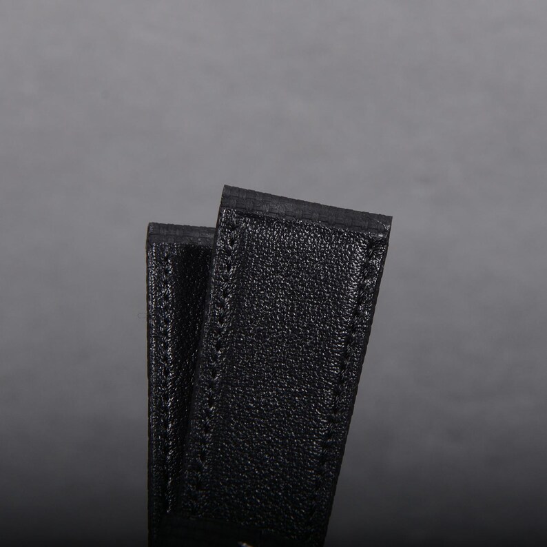 May include: Close-up of two black leather straps, possibly for a watch or bag. The straps have a textured surface and are stitched along the edges. The background is a solid gray.