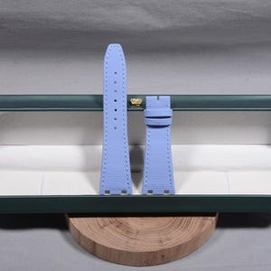 Puede incluir: Una correa de reloj azul claro con costuras blancas. La correa est&aacute; hecha de un material texturizado y tiene un cierre de hebilla. La correa se muestra en una caja verde abierta con un emblema dorado.