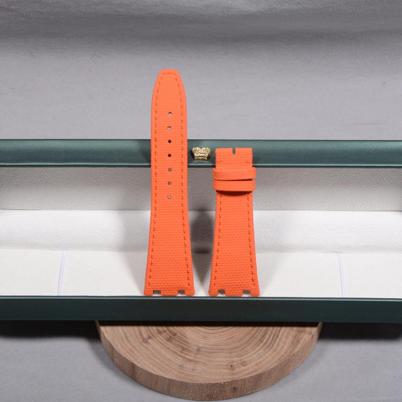 Puede incluir: Juego de correas de reloj de color naranja brillante. Las correas est&aacute;n hechas de un material texturizado y presentan costuras en contraste. El juego incluye dos piezas, una con agujeros para la hebilla y otra con una hebilla. Las correas se muestran en una caja verde.