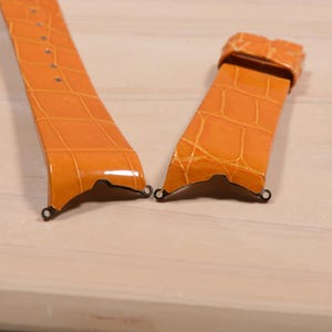 May include: Two-piece orange watch strap with a crocodile skin texture. The strap has a glossy finish and metal hardware for attachment. The watch strap is shown on a light wood surface.