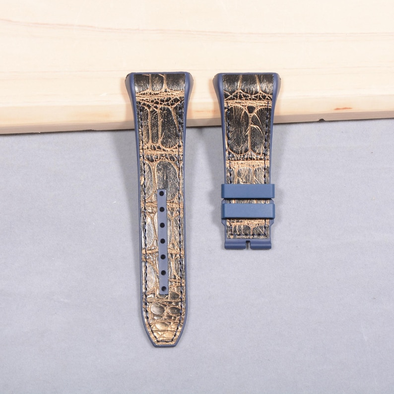 K&ouml;nnte beinhalten: Zweiteiliges Uhrenarmband-Set. Die Armb&auml;nder sind blau mit einem gold-braunen Krokodilmuster. Das linke Armband hat mehrere L&ouml;cher zur Anpassung. Das rechte Armband hat eine blaue Schnalle.