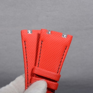 May include: Close-up of two bright red watch straps with a textured weave pattern. The straps have black stitching and silver metal clasps. The watch straps are shown against a gray background.