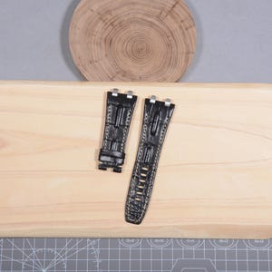 May include: Two black watch straps with a crocodile pattern and silver-tone hardware are displayed on a wooden surface. The straps are made of leather and have contrasting stitching. The background includes a wooden slice and a gray surface.