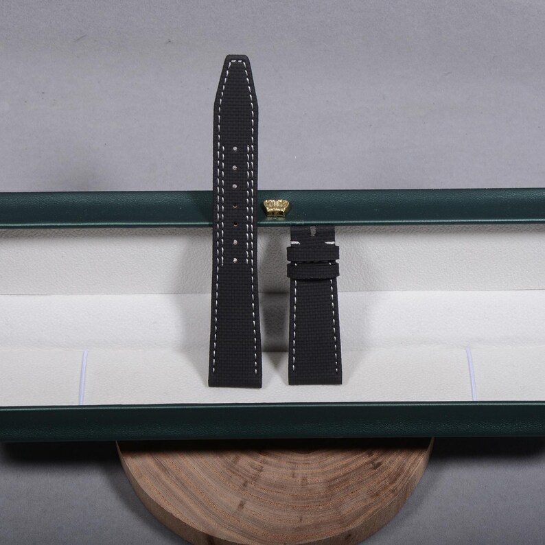 May include: Black watch strap with white stitching, displayed in an open green box with a white interior. The strap is made of a textured material. The box has a gold-colored crown-shaped accent.