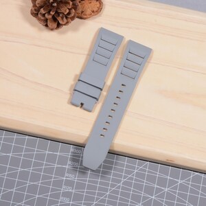 Op de afbeelding: Twee grijze horlogebandjes liggen op een houten oppervlak. De bandjes zijn gemaakt van een flexibel materiaal en hebben rechthoekige inkepingen en verstelgaten. De bandjes zijn ontworpen om aan een horlogekast te bevestigen.