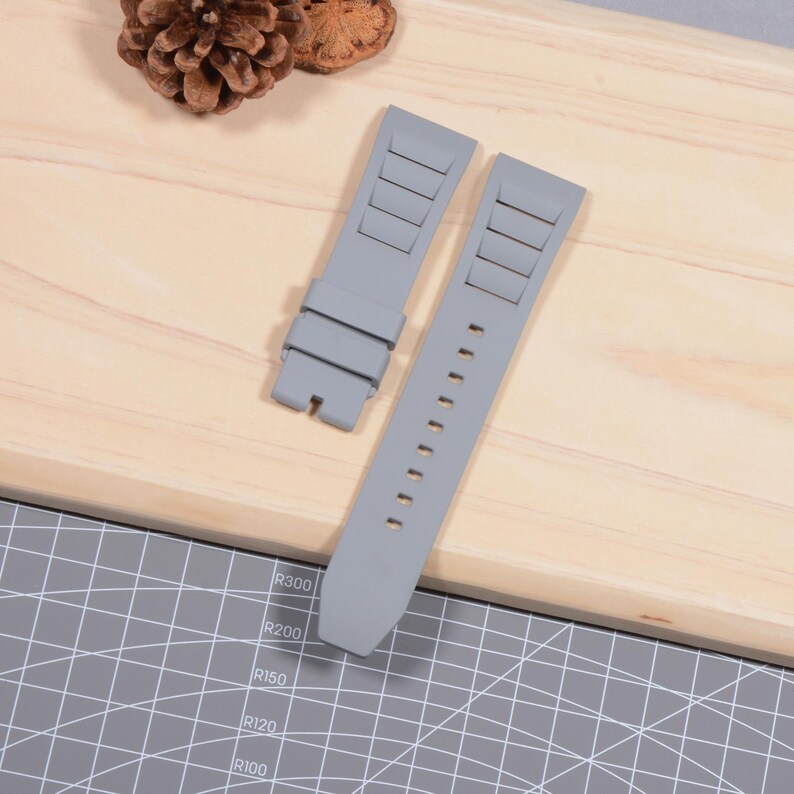 Op de afbeelding: Twee grijze horlogebandjes liggen op een houten oppervlak. De bandjes zijn gemaakt van een flexibel materiaal en hebben rechthoekige inkepingen en verstelgaten. De bandjes zijn ontworpen om aan een horlogekast te bevestigen.