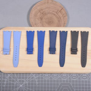 Puede incluir: Surtido de correas de reloj en varios tonos de azul y gris, expuestas sobre una tabla de madera. Las correas son de material texturizado y presentan costuras y orificios para su ajuste. Una rodaja de madera se encuentra en el fondo.