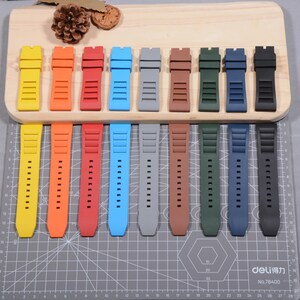 Op de afbeelding: Een verzameling kleurrijke horlogebandjes in verschillende tinten, waaronder geel, oranje, rood, blauw, grijs, bruin, groen, marineblauw en zwart. De bandjes zijn gemaakt van flexibel materiaal en worden getoond op een houten plank en een grijze snijmat.