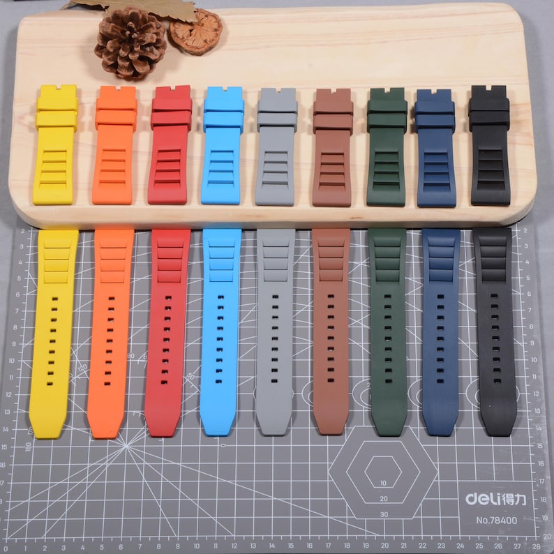 Op de afbeelding: Een verzameling kleurrijke horlogebandjes in verschillende tinten, waaronder geel, oranje, rood, blauw, grijs, bruin, groen, marineblauw en zwart. De bandjes zijn gemaakt van flexibel materiaal en worden getoond op een houten plank en een grijze snijmat.