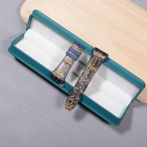 K&ouml;nnte beinhalten: Zwei Uhrenarmb&auml;nder mit Krokodilleder-Muster in einer teal-farbenen Box. Die Armb&auml;nder sind braun und goldfarben mit blauen Akzenten. Die Box ist teal mit wei&szlig;em Innenraum und liegt auf einer grauen Oberfl&auml;che.