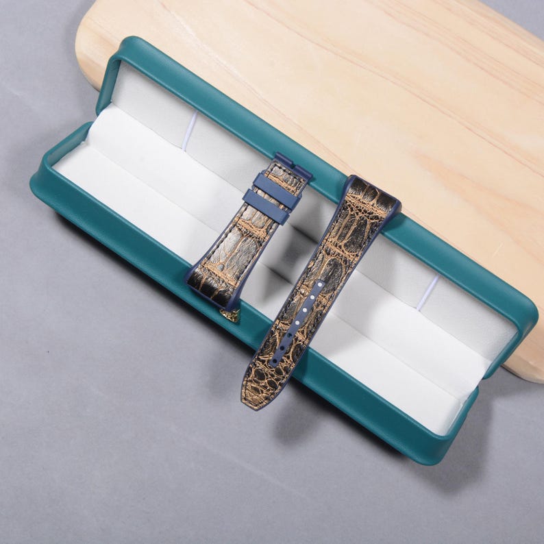 K&ouml;nnte beinhalten: Zwei Uhrenarmb&auml;nder mit Krokodilleder-Muster in einer teal-farbenen Box. Die Armb&auml;nder sind braun und goldfarben mit blauen Akzenten. Die Box ist teal mit wei&szlig;em Innenraum und liegt auf einer grauen Oberfl&auml;che.