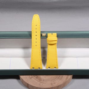 Puede incluir: Correa de reloj amarilla de dos piezas con superficie texturizada. La correa tiene un borde cosido y una serie de agujeros para ajustar. La correa del reloj se muestra dentro de una caja verde y blanca.