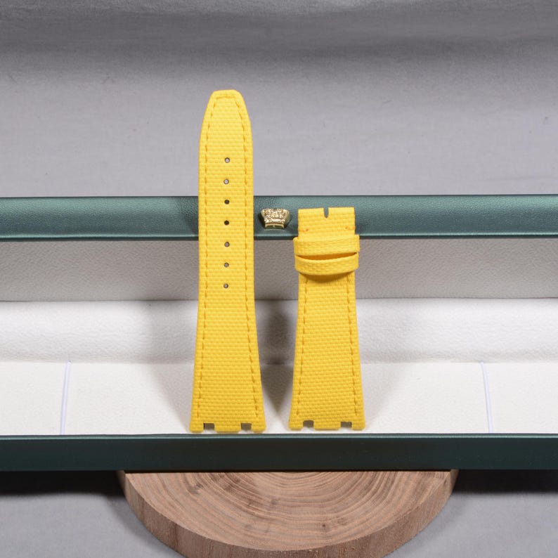 Puede incluir: Correa de reloj amarilla de dos piezas con superficie texturizada. La correa tiene un borde cosido y una serie de agujeros para ajustar. La correa del reloj se muestra dentro de una caja verde y blanca.