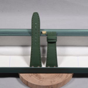 Puede incluir: Un par de correas de reloj verde oscuro con costuras en contraste. Las correas est&aacute;n hechas de un material texturizado y se exhiben dentro de una caja verde con un emblema dorado. Las correas descansan sobre una base de madera.