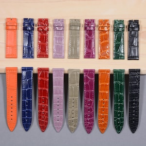 May include: A collection of colorful watch straps in various shades, including orange, blue, red, purple, and green. The straps have a textured, crocodile-like pattern and are displayed on a wooden surface. Each strap is designed to fit a watch.