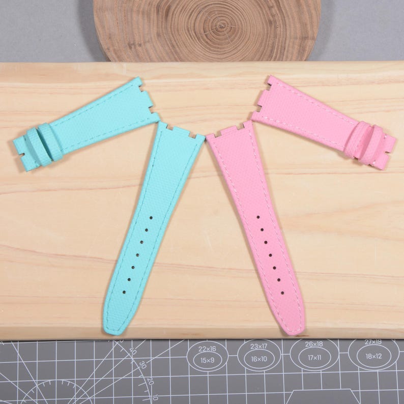 Puede incluir: Dos correas de reloj, una en azul claro y otra en rosa, se muestran sobre una superficie de madera. Cada correa tiene una superficie texturizada, agujeros de ajuste y una hebilla. Las correas est&aacute;n dise&ntilde;adas para un reloj.