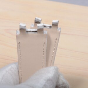 May include: Close-up of two light beige leather watch straps with white metal clasps. The straps are stitched with light thread and held by a gloved hand. The background is a light wood surface.