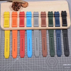 Op de afbeelding: Een verzameling horlogebandjes in verschillende kleuren, waaronder geel, oranje, rood, blauw, grijs, bruin, groen, marineblauw en zwart. De bandjes zijn gemaakt van flexibel materiaal en hebben rechthoekige uitsparingen en kleine gaatjes.