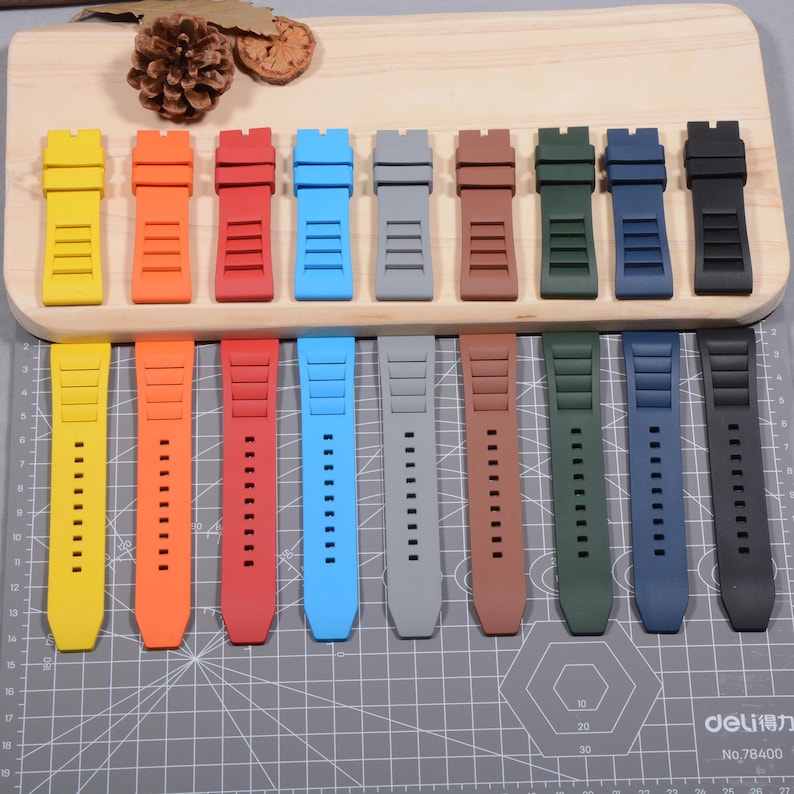 Op de afbeelding: Een verzameling horlogebandjes in verschillende kleuren, waaronder geel, oranje, rood, blauw, grijs, bruin, groen, marineblauw en zwart. De bandjes zijn gemaakt van flexibel materiaal en hebben rechthoekige uitsparingen en kleine gaatjes.