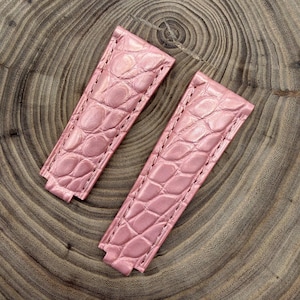 Könnte beinhalten: Zwei rosa Uhrenarmbänder mit Krokodilleder-Textur. Die Armbänder sind aus Leder gefertigt und haben ein genähtes Design. Die Armbänder werden auf einer Holzoberfläche gezeigt.