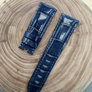 Puede incluir: Correa de reloj azul marino de dos piezas con un patrón de cocodrilo. La correa de cuero tiene costuras azul claro y un acabado pulido. La correa se muestra sobre una superficie de madera.