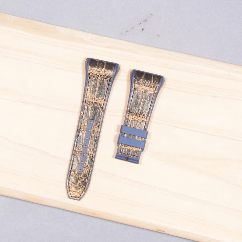 K&ouml;nnte beinhalten: Zwei Uhrenarmb&auml;nder mit Krokodilmuster in Braun- und Graut&ouml;nen, mit blauen R&auml;ndern und Akzenten. Die Armb&auml;nder werden auf einer hellen Holzoberfl&auml;che pr&auml;sentiert.