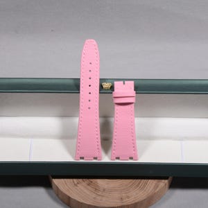 Puede incluir: Correa de reloj rosa de dos piezas con superficie texturizada. La correa tiene un borde cosido y varios agujeros para ajuste. La correa se muestra dentro de una caja verde con un emblema de corona dorada.