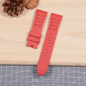 Op de afbeelding: Tweedelig rode horlogeband. De band is gemaakt van een flexibel materiaal en heeft rechthoekige uitsparingen aan &eacute;&eacute;n kant. De andere kant heeft meerdere gaten voor aanpassing. De band wordt getoond op een licht houten oppervlak.